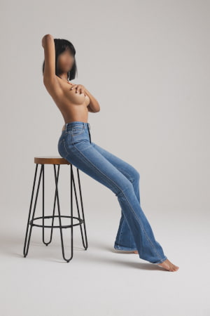 Topless exotic woman in flared jeans sitting on a stool in an empty room